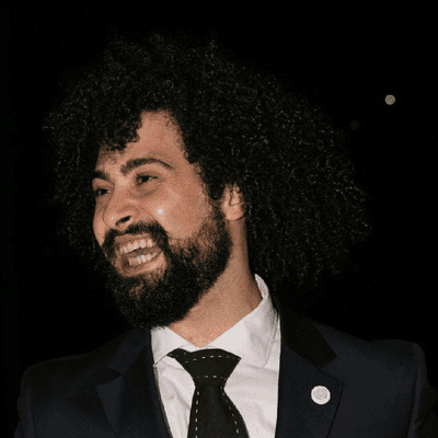 Claudio Longhitano, fa tante cose Ochianu a Calascibetta. Questo è un ritratto fotografico notturno di Claudio, che appare con i suoi distintivi capelli ricci e scuri e una barba stranamente ben curata (per una volta). Claudio è colto a mezza altezza e guarda fuori campo a sinistra con un ampio e sincero sorriso. Indossa un abito sartoriale blu scuro, con una spilla rotonda di MSOI—Movimento Studentesco per l’Organizzazione Internazionale. La sua camicia è bianca pulita e la cravatta scura presenta un motivo geometrico a puntini. Lo sfondo è nero scuro e sfocato, con un piccolo e luminoso bokeh in alto a destra.